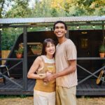 Pareja sonriente brindando con copas cerca de una caravana.